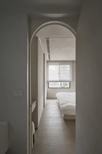 A minimalist bedroom featuring light-colored wood flooring, white bedding, and a large window with blinds.