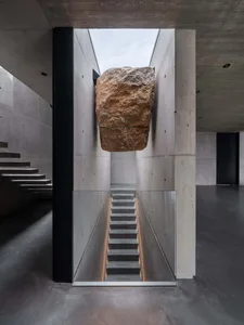 A minimalist stairwell with concrete walls and flooring, featuring a large rock embedded in the ceiling.