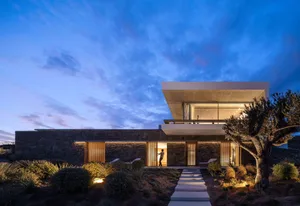 Modern residential home with concrete exterior, wooden windows, and glass doors, illuminated at dusk with blue sky.
