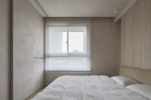 A minimalist bedroom with neutral-toned walls, a bed with white bedding, and white blinds on the window.