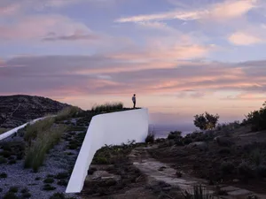 A person standing on a rooftop wall overlooking a sunset and distant sea.