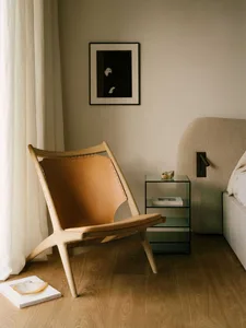 A minimalist bedroom featuring a wooden frame, leather upholstered lounge chair, and a glass top bedside table.