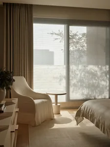 A minimalist white bedroom featuring a sleek white chair, a round white side table, and a large window with blinds, overlooki