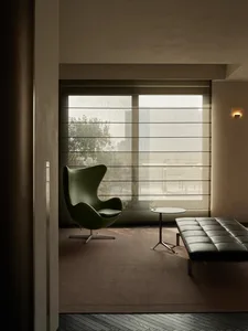 A modern interior space featuring a green egg chair, round side table, black leather bench, and large window with beige blind