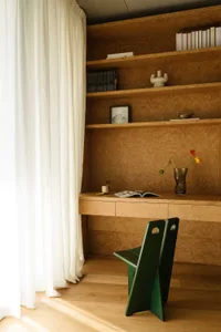 A modern, minimalist home office with wooden furniture, white curtains, and a green chair.