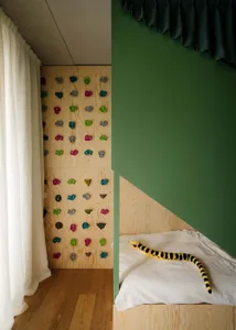 A minimalist playroom featuring a wooden climbing wall with colorful grips and a bed with a snake-shaped plush toy.