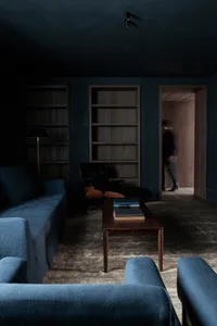 Minimalist residential interior with concrete walls, wooden shelves, leather armchair, and blue sofas.