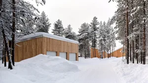 A modern winter cabin with wooden exterior and white roofs amidst a snowy, forested landscape.