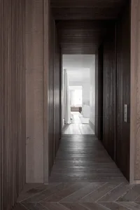 A modern wooden corridor with dark wooden floors and walls, leading to a bright and airy room with white furniture and curtai