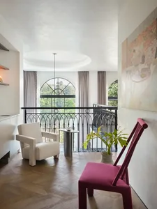 A modern living room with wooden furniture, a white armchair, a pink chair, and a large arched window.