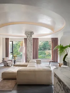 A modern living room featuring beige leather furniture, large windows with pink curtains, and a marble column and floor.