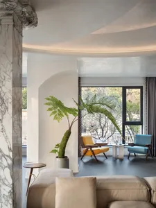 A modern minimalist living room with concrete walls, a beige sofa, a yellow chair, a blue chair, and a potted fern plant.