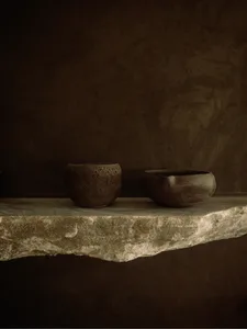 Two ceramic bowls made of clay material, placed on a stone shelf against a dark brown wall.