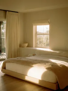 Minimalist bedroom with textured fabrics, wooden floor, and natural light streaming in through curtains and windows.
