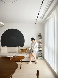 A minimalist living room with a beige sectional sofa, wooden tables, and a black circular accent on the wall.