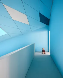 A minimalist interior with blue walls, geometric ceiling tiles, and a baby sitting on the floor.