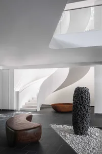 A minimalist lobby with concrete walls and floor, featuring a curved wooden bench, a dark textured pillar, and a large bowl-s