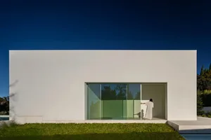 Modern residential exterior with a minimalist design, featuring a white plaster facade and large glass windows.