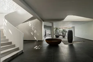 Modern lounge area with concrete walls and floor, featuring a spiral staircase, a large round metal planter, and greenery in 
