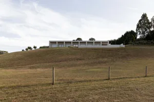 A sleek, modern house with concrete and glass materials sits atop a grassy hillside.