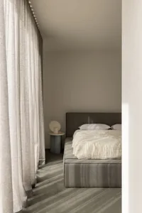 A modern bedroom featuring a grey upholstered bed with white bedding, surrounded by sheer curtains and a textured grey carpet