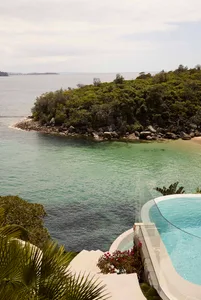 A modern waterfront home with a glass pool overlooking the ocean and lush greenery.