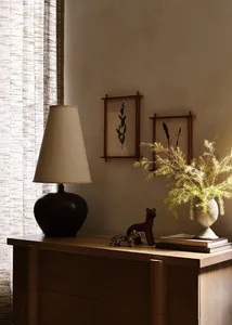 A minimalist interior design featuring a wooden dresser with ceramic vase and lamp, and framed botanical prints on the wall.