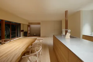A minimalist residential kitchen with wooden countertops, flooring, and chairs, featuring a large kitchen island with a sink.