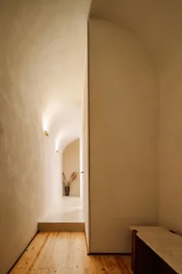 A minimalistic hallway with concrete walls and a wooden floor, featuring a vase with dried plants.
