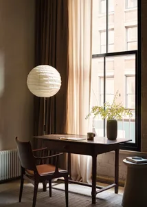 A modern home office featuring a wooden desk with a white paper lantern lamp, brown curtains, and a vase with flowers.