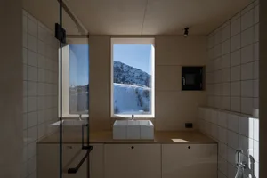 A minimalist bathroom featuring white tiles and wood elements, with a view of snowy mountains through a window.