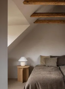 A minimalist bedroom with wooden ceiling beams and a bed covered in grey fabric, featuring a small wooden side table with a w