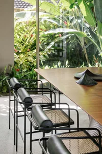 Modern minimalist dining table with black metal frame and woven rattan seating surrounded by lush greenery through large wind