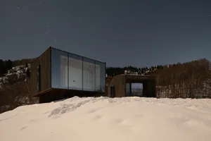 Modern residential architecture with glass and wood exterior at night in a snowy landscape.