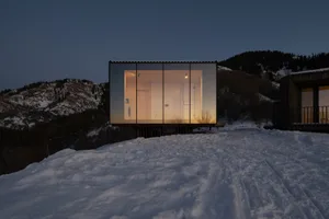 極簡石屋搭配玻璃牆與木質露台，俯瞰黃昏時分的雪丘景觀。