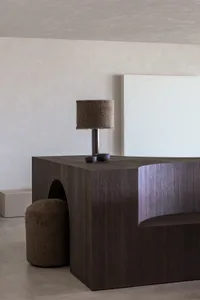 A modern interior design featuring a wooden table with a lamp, a white wall with a circular pattern, and a plush ottoman.