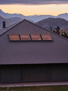 The exterior of a modern house featuring a metal roof with three square windows at sunset with mountainous scenery in the bac