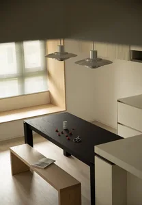 A modern residential kitchen with wooden accents, featuring a black dining table, a white bench, and a window with blinds.