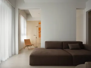 A minimalist living room featuring a brown sectional sofa with textured fabric, a tall lamp, and a window with sheer curtains