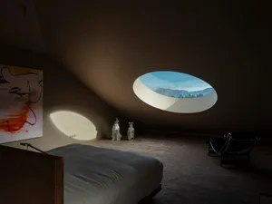 A minimalist bedroom featuring a concrete floor, white bed, and a circular window overlooking mountains.