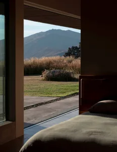 A modern residential bedroom with large windows overlooking grassy fields and distant mountains.