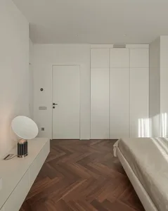 Modern minimalist bedroom with white walls, wooden flooring, and a single white lamp on a floating nightstand.