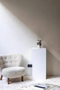 A minimalist living room with a sheepskin armchair, a metallic vase on a pedestal, and an open book on a stone table with a m
