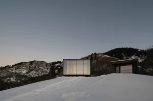 A modern, rectangular house with large glass windows on a snowy hillside.