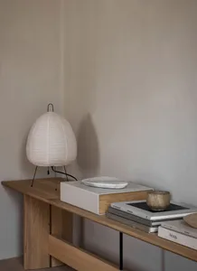 A modern living room with a wooden table, a white lampshade, and some books on a wooden shelf.