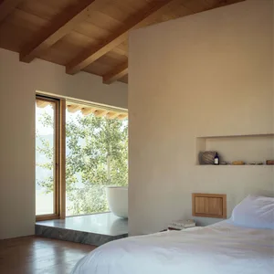 A bedroom with minimalist design elements, featuring a concrete floor, wooden beams, and a bathtub with a view of the wooded