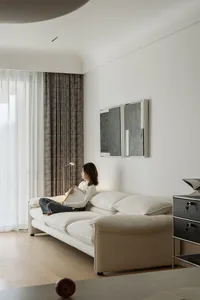 A modern minimalist living room features a white couch with plush cushions and a large window with sheer drapes, allowing nat