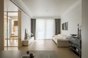 A modern living room with neutral-toned furniture and minimalistic decor, featuring a large window with sheer curtains allowi