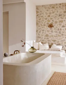 A minimalist bathroom featuring a concrete tub and a cozy nook with a stone wall and white cushions.