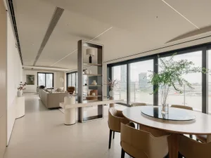 A modern, neutral-toned living space featuring a dining table and chairs, a round table, a long sofa, and tall windows with a